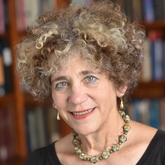 Headshot of Johanna Drucker wearing a black top and necklace against a bookshelf. Image courtesy of Johanna Drucker.